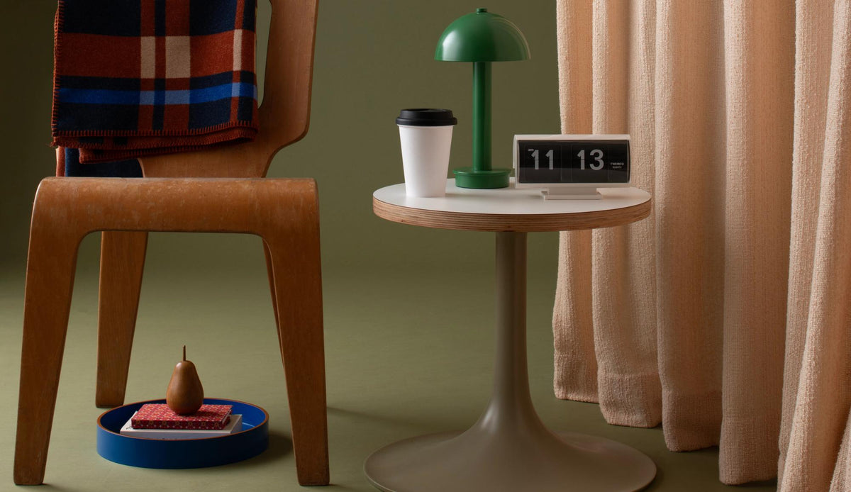 Organized minimalist gift guide scene with a green lamp and round side table displaying a flip clock, cup, chair, and tray