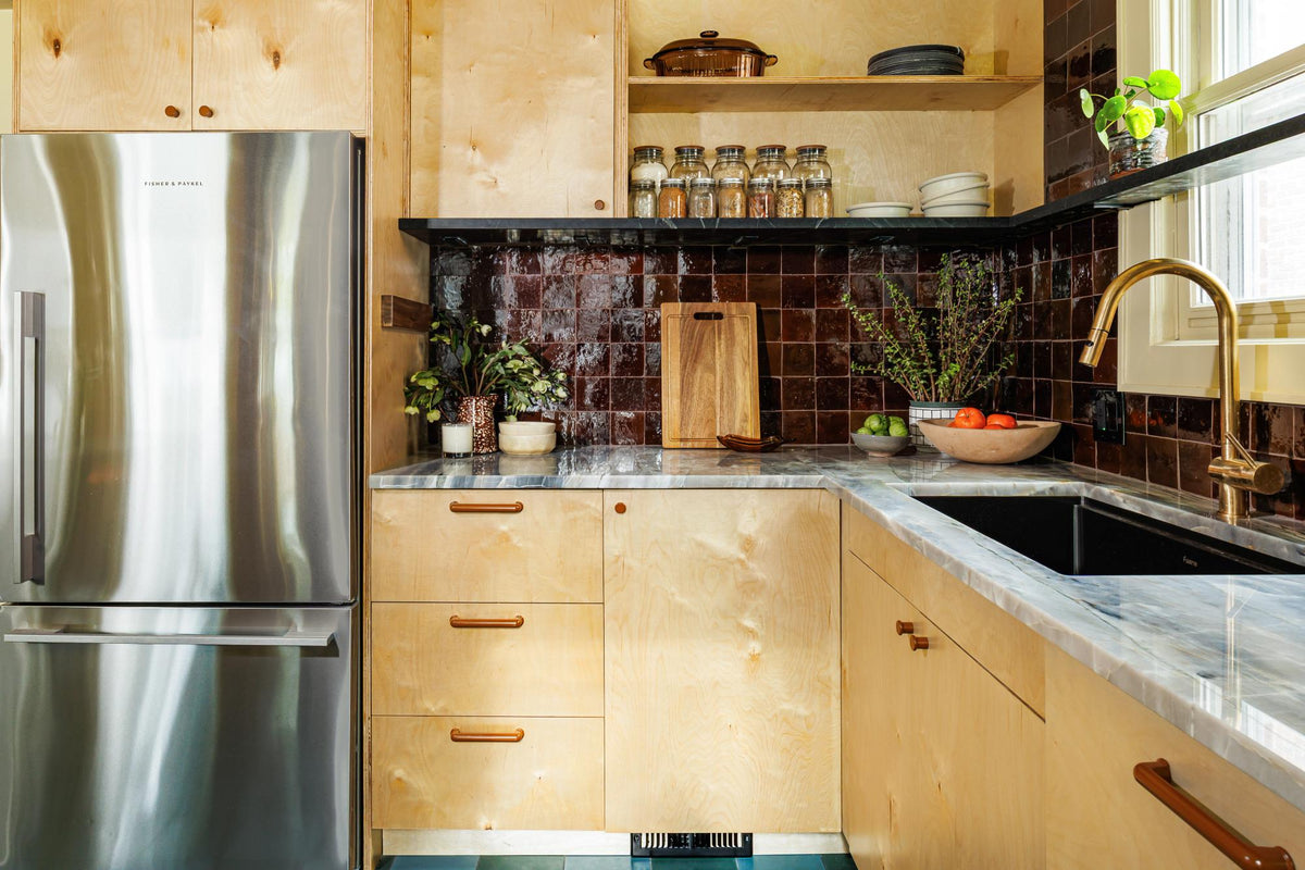 Eclectic kitchen features wood cabinets, dark tile backsplash, marble counters, brass hardware, and a stainless steel refrigerator