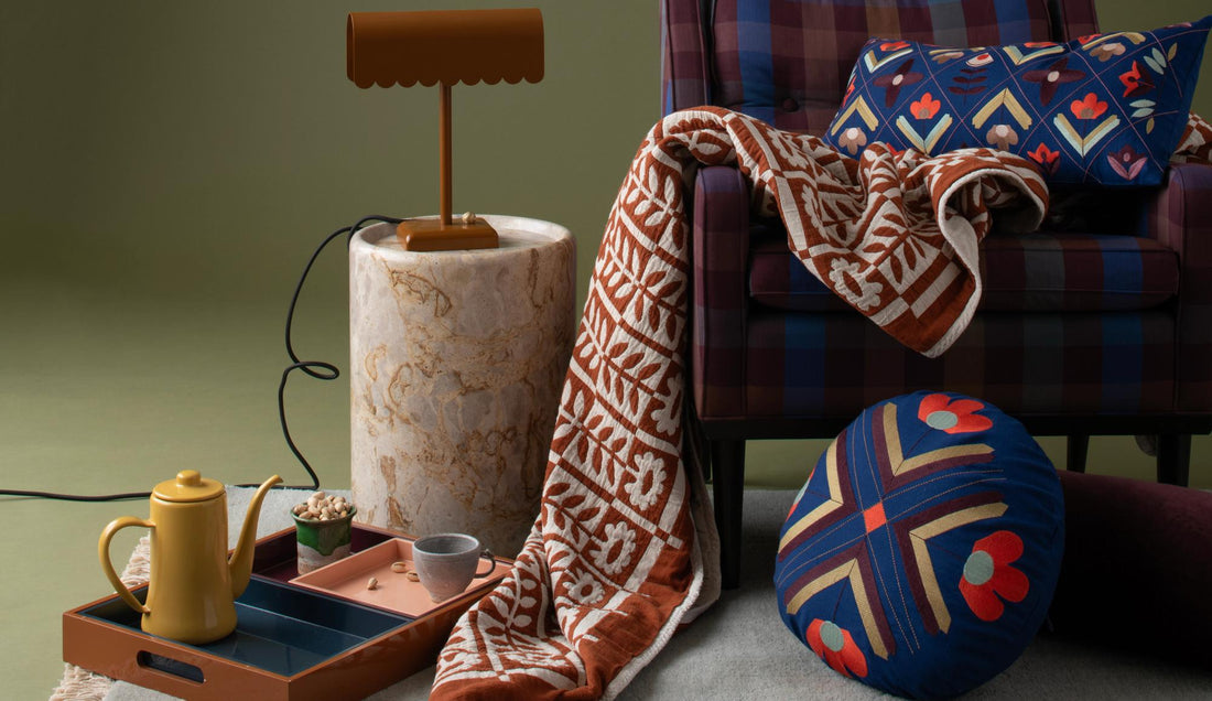 Cozy seating area with a plaid chair, patterned pillows, draped quilt, lamp, tray, and tea set