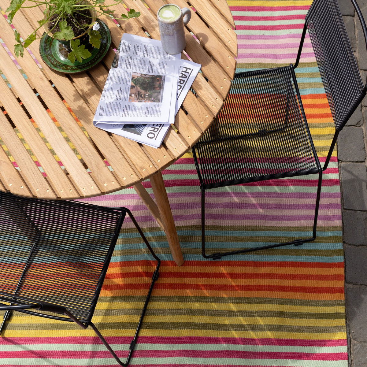 Outdoor table and chairs arranged on a colorful striped rug with a plant, coffee, and newspaper