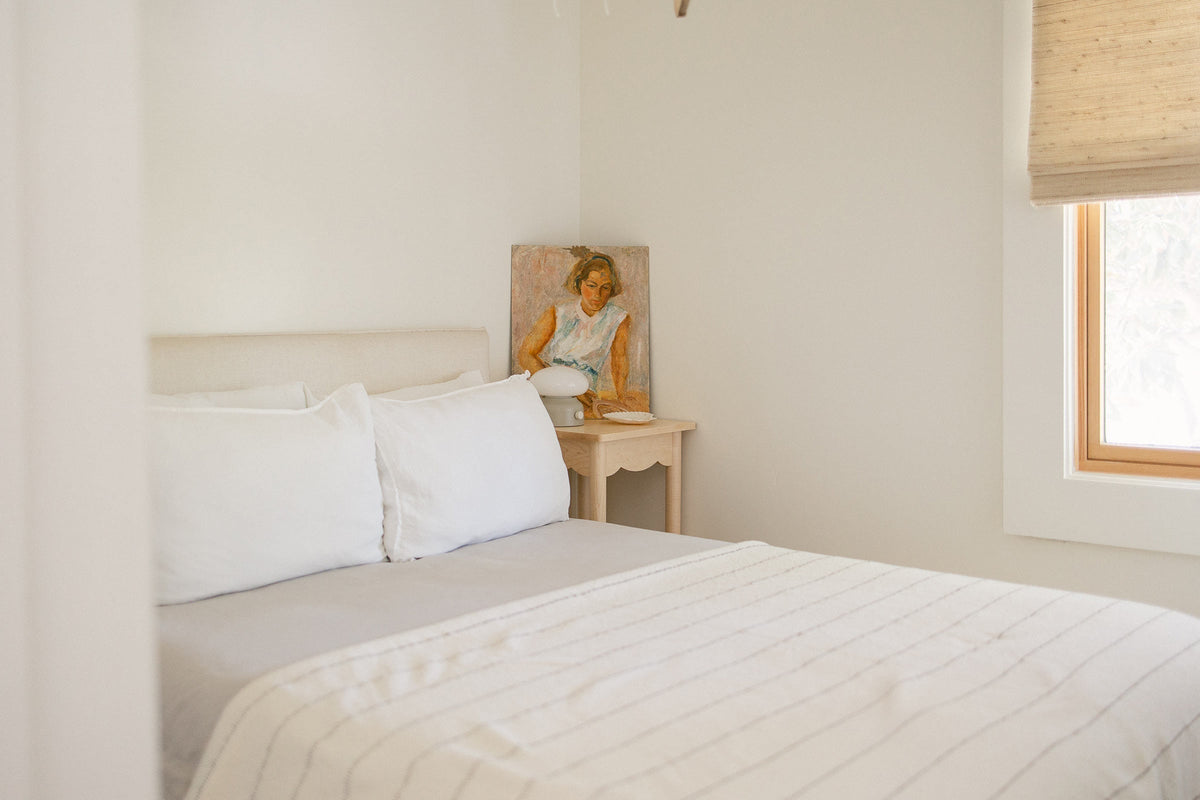 Neutral bedroom with made bed, white pillows and striped coverlet, nightstand with lamp and leaning portrait, soft window light