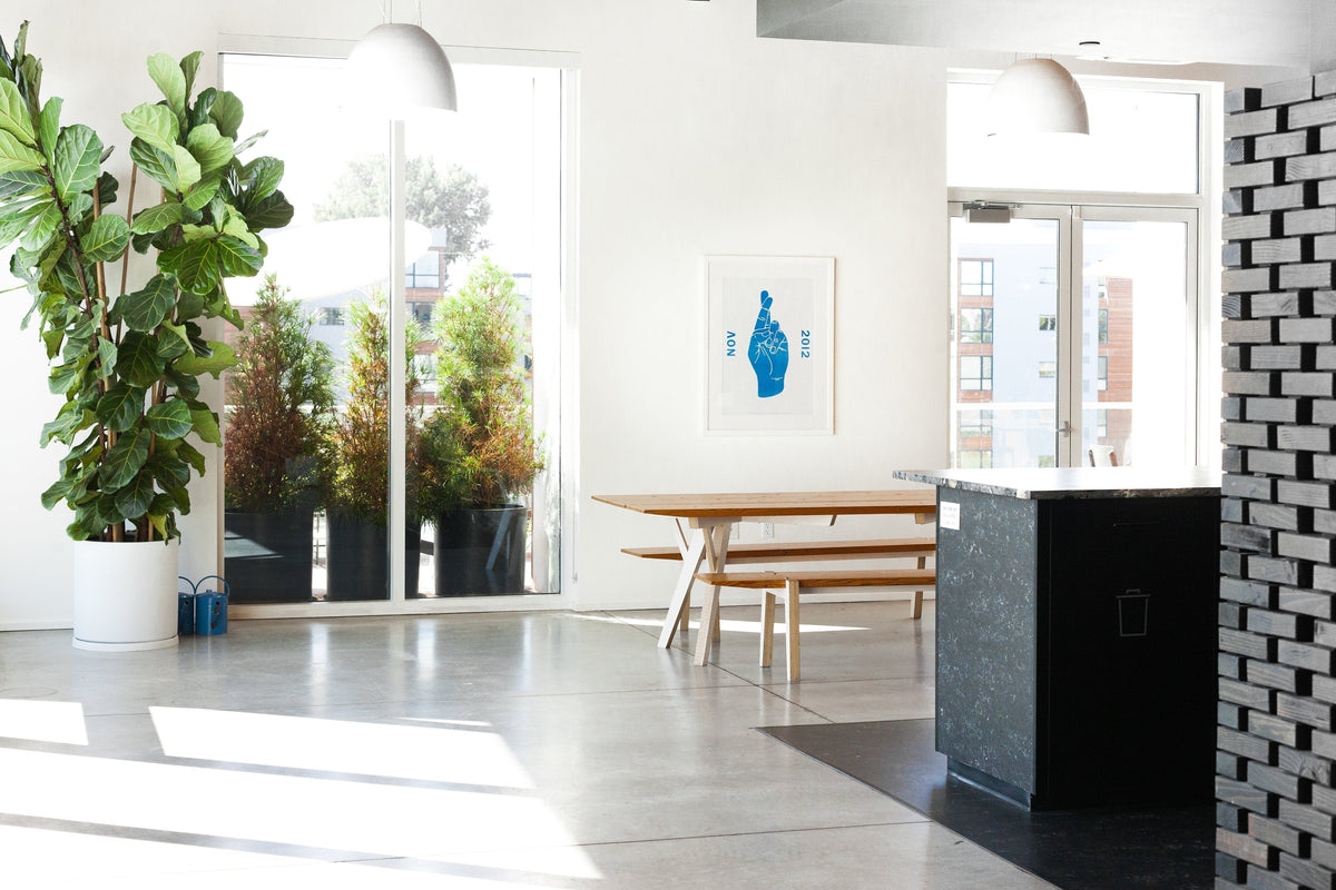Sunlit dining space with picnic-style table and bench, fiddle-leaf fig by tall windows, framed print reading NOV 2012