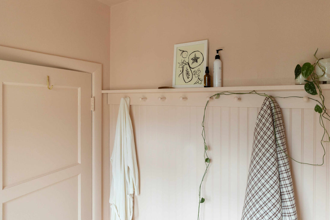 Pale pink beadboard wall in an entryway with a peg rail, a white robe and plaid towel hanging, framed artwork and a trailing pothos plant on a narrow shelf
