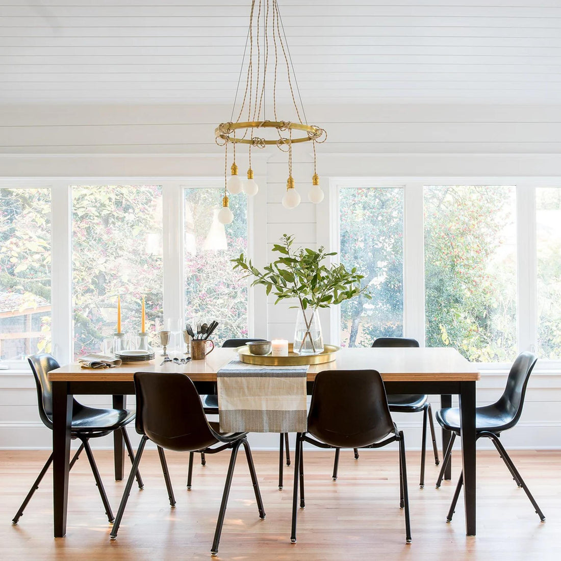 Dining table set with dishes, candles, and a leafy centerpiece in a bright room with large windows