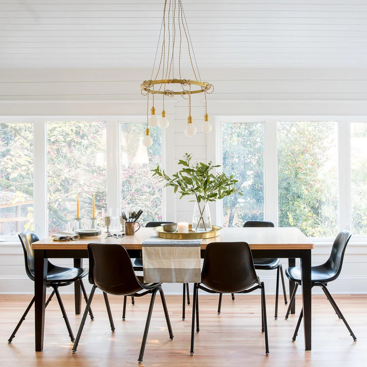 Dining table set with dishes, candles, and a leafy centerpiece in a bright room with large windows