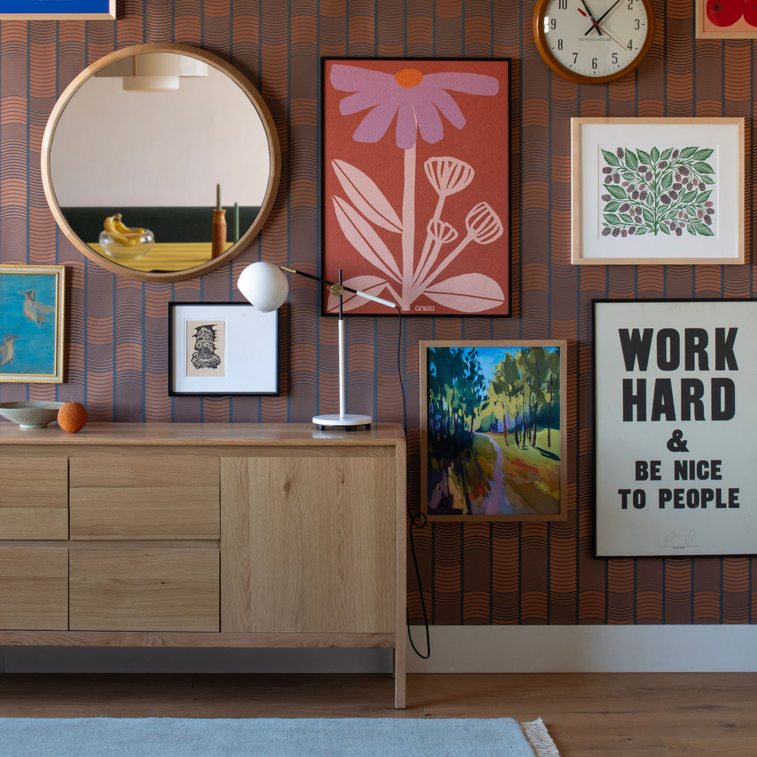 Maximalist gallery wall with framed artwork, a round mirror, clock, lamp, and cabinet in a patterned room