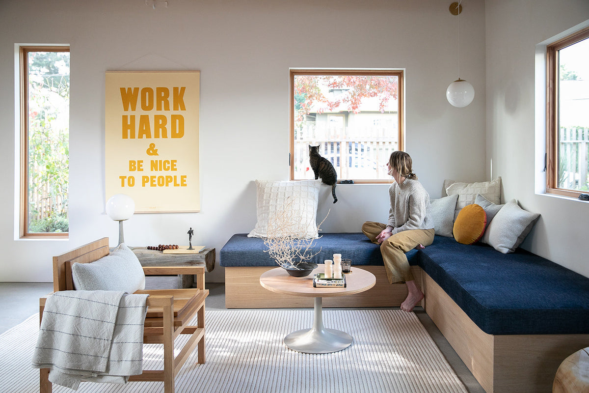 Woman on bench watching cat on windowsill in living room, yellow poster reads WORK HARD AND BE NICE TO PEOPLE
