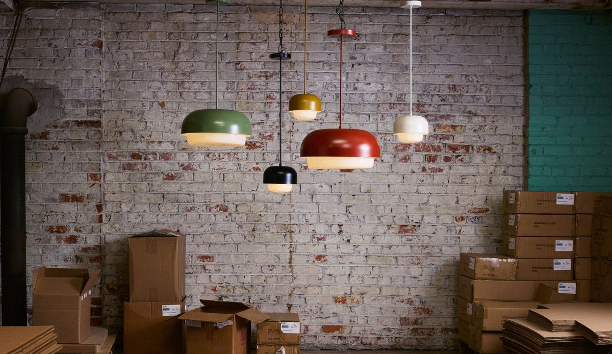 Six colorful pendant lamps from the Benchmade Ray Collection hang against a brick wall in a workshop