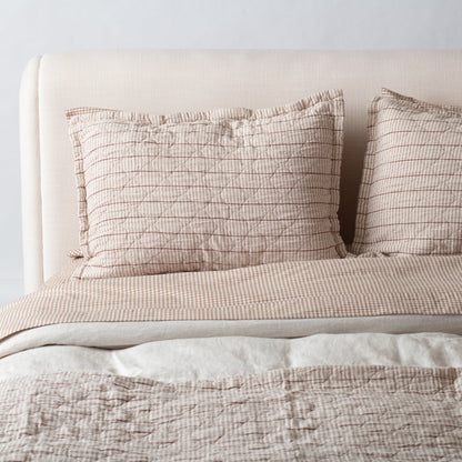 Front view of two Diamond Ticking Quilted Pillow Shams in russett linen with thin stripes and diamond quilting on a cream upholstered headboard