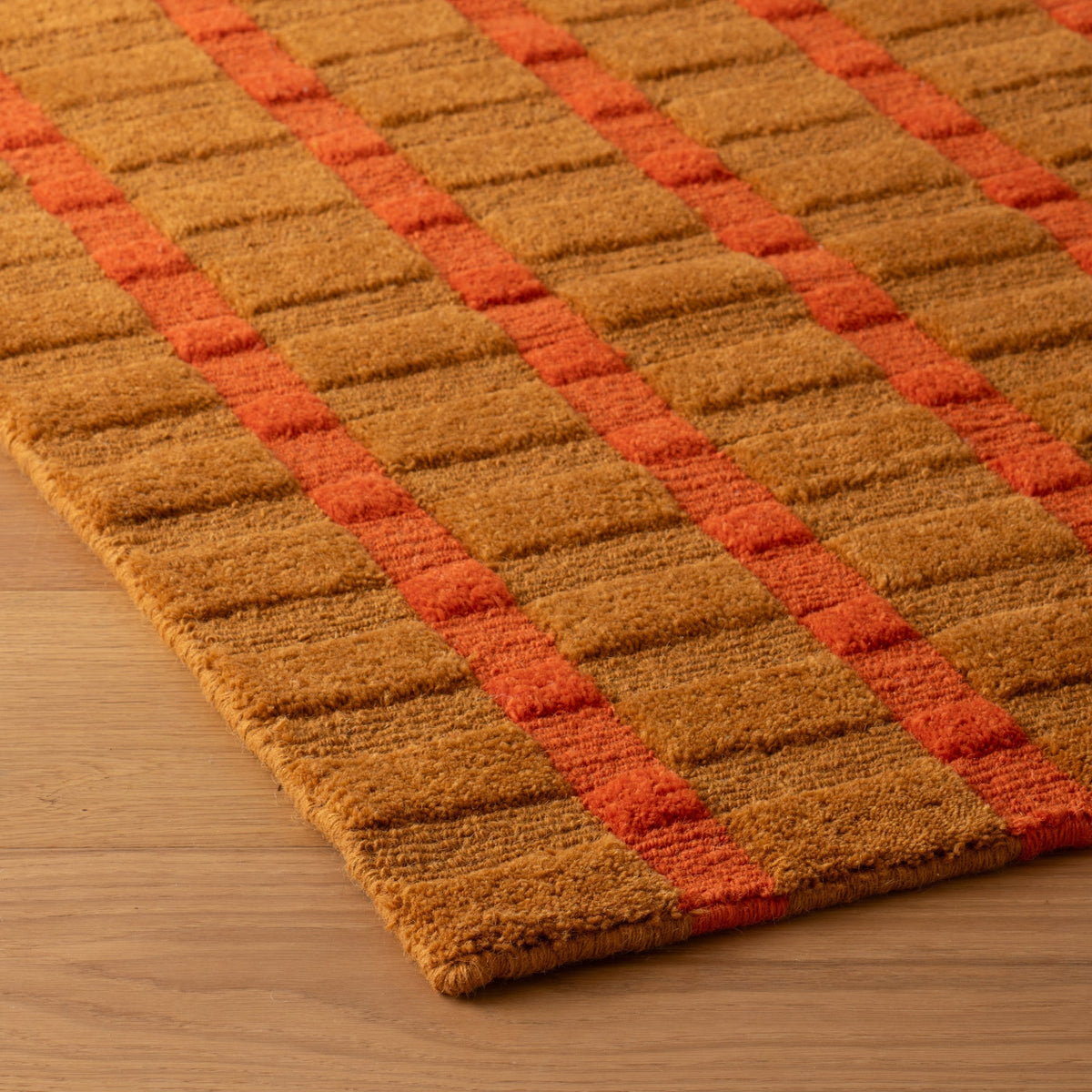 Angled view of Ashlar Rug showcasing a raised golden brown grid with orange red stripes on a wood floor