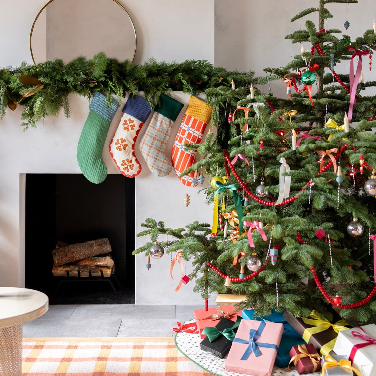 Holiday stockings hang from a greenery-covered mantel beside a decorated Christmas tree and wrapped gifts