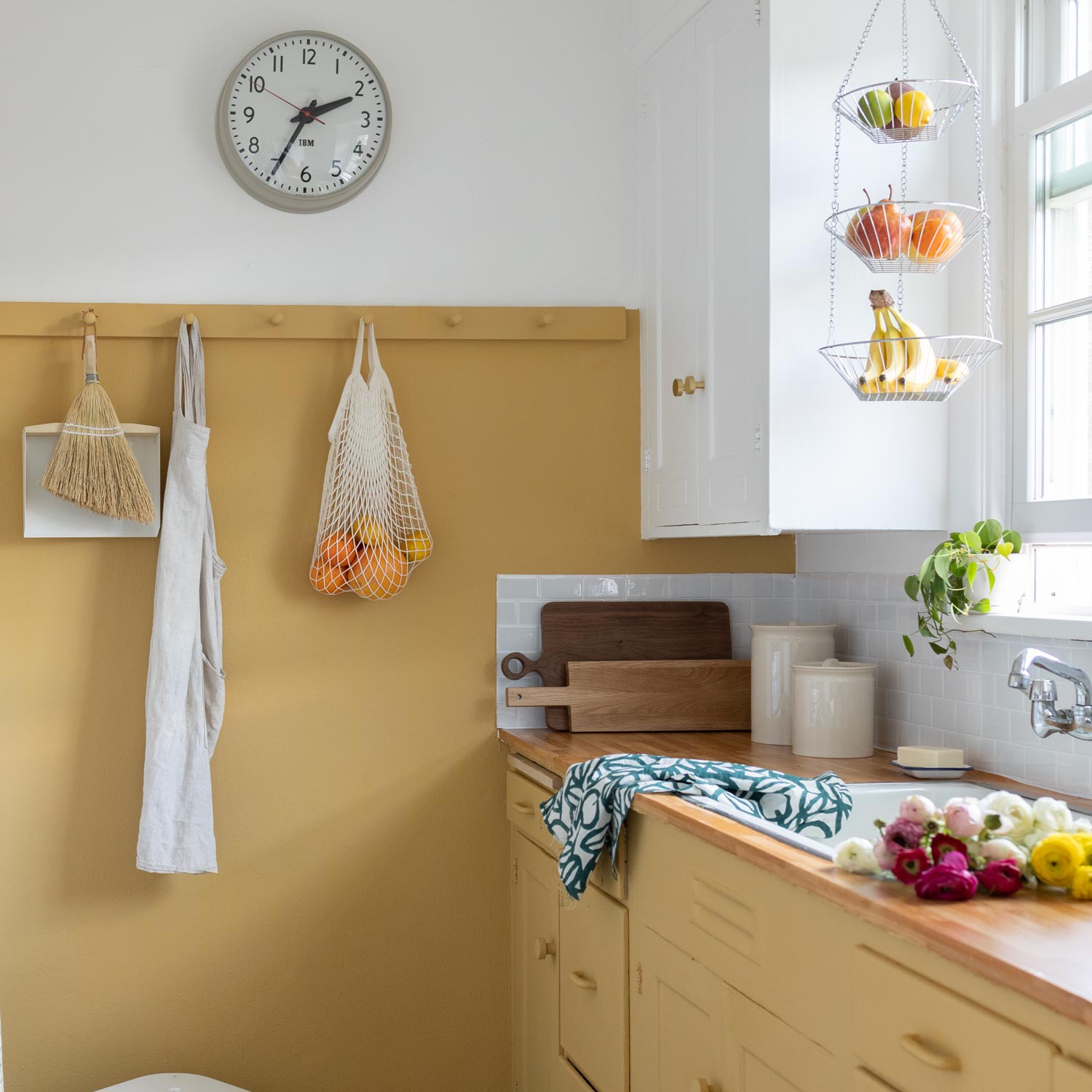 Bright kitchen with mustard cabinets, wood countertop, sink, wall hooks, hanging produce baskets, and flowers