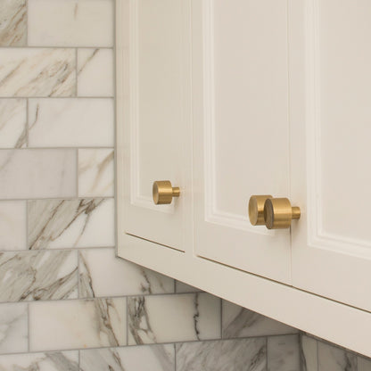 Angled view of Mid Century Knob showcasing cylindrical hardware on cream cabinet doors with marble tile background
