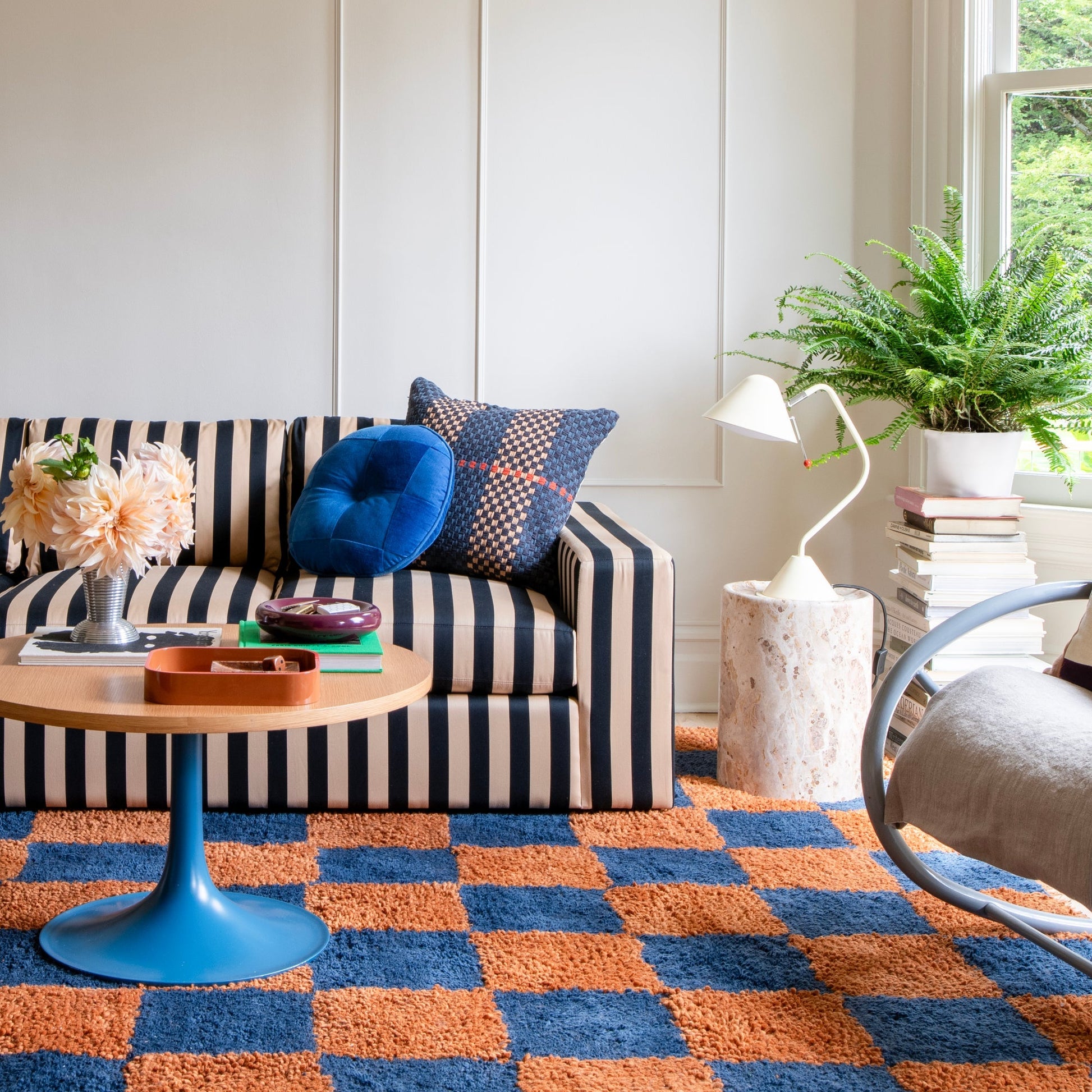 Jacques shag wool rug displayed under a striped sofa in a bright living room with blue pillows, a potted fern, and a white table lamp