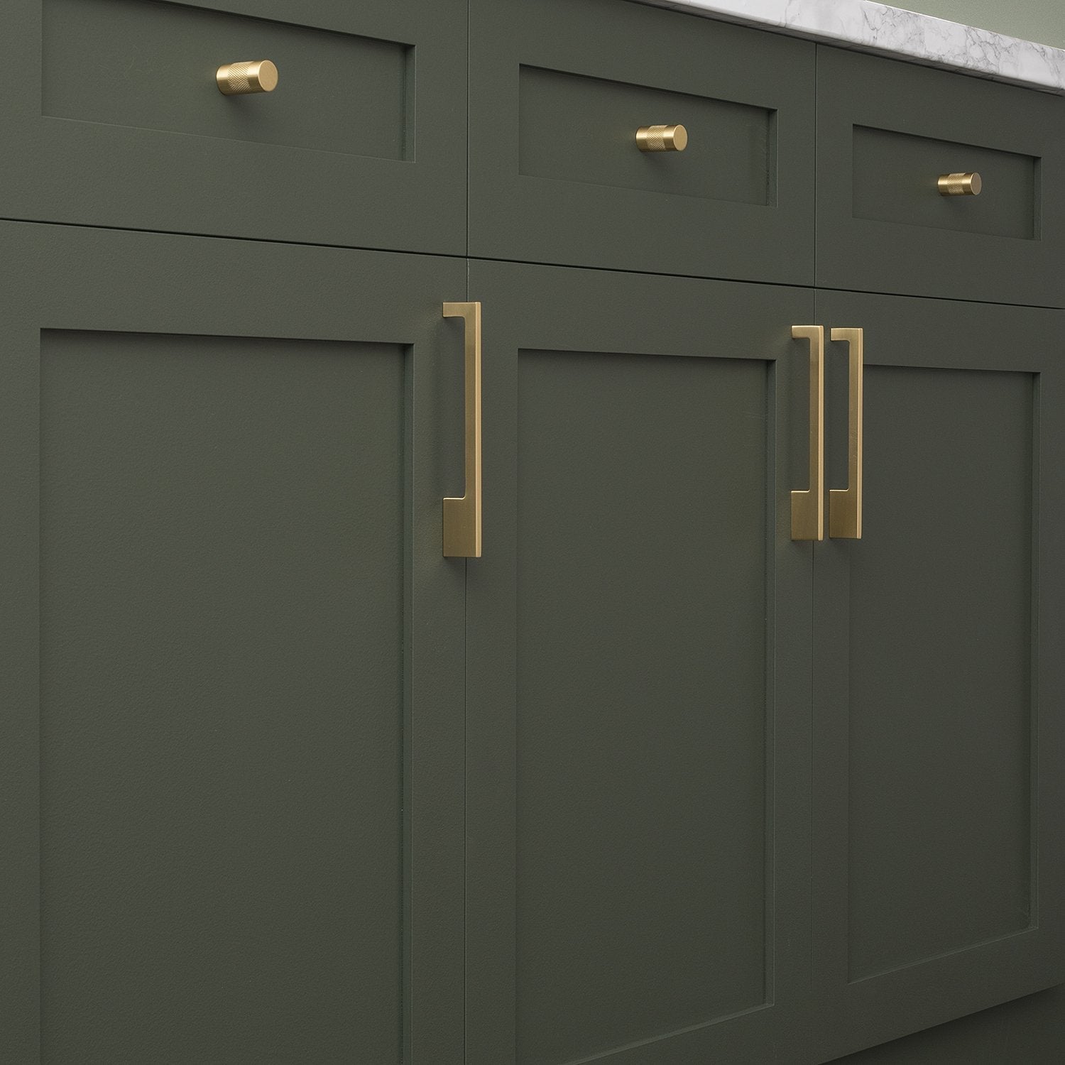 Natural brass knurled knobs adorn dark green kitchen cabinets beneath a marble countertop