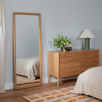 Oak six-drawer dresser displayed in a bedroom with a mirror, lamp, flowers, and beds