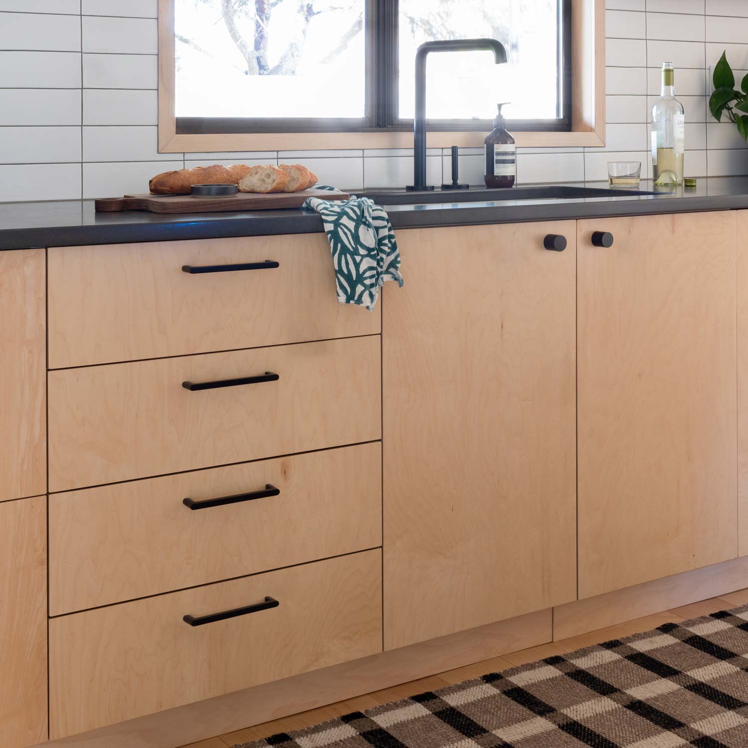 Black Cove Pulls installed on light wood kitchen cabinets beside a sink and tiled backsplash