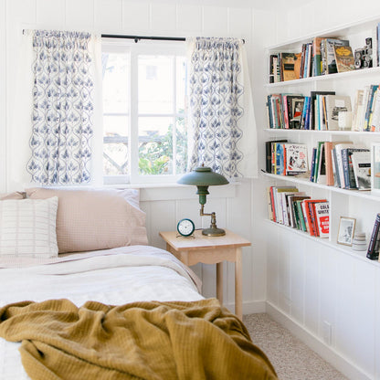 Bed with russet gingham pillowcases beside a wooden nightstand and bookshelf in a bright cozy bedroom